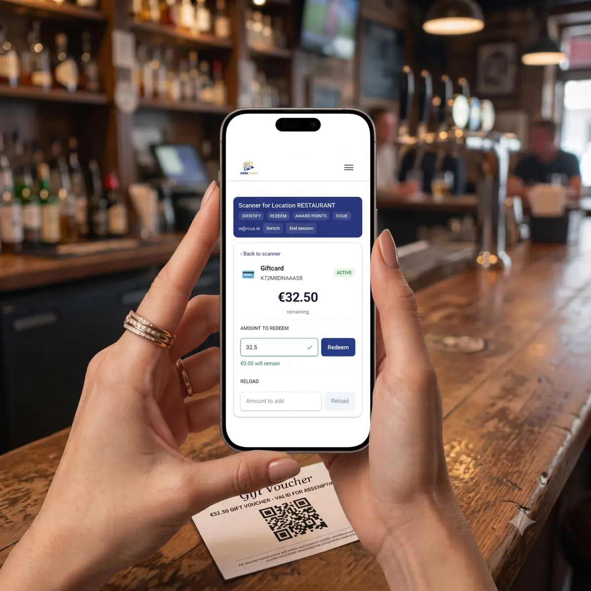 Staff at a bar counter holding a phone showing the Keyring scanner after a scan: the venue name and its capabilities across the top, then the instrument identified as a gift card with its code, an active status, the balance remaining, and an amount field beside a redeem button. A printed voucher carrying a QR code lies on the counter.